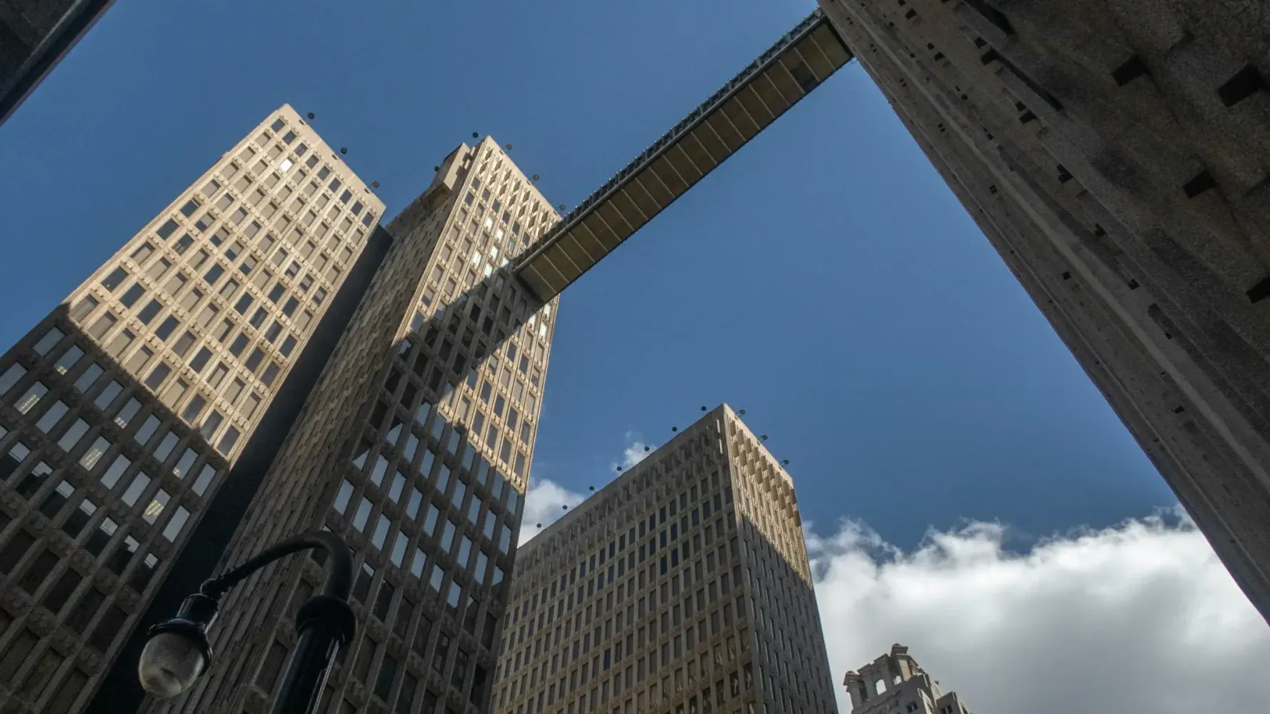 Two tall office towers connected by a skybridge, viewed from below with blue sky and clouds above.