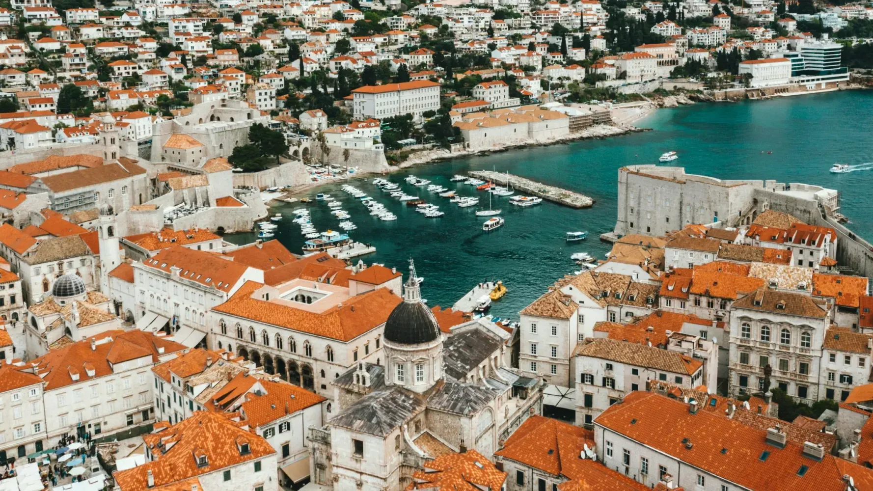 Aerial view of Dubrovnik’s old harbor with yachts, medieval walls, and historic stone buildings with red-tiled roofs.