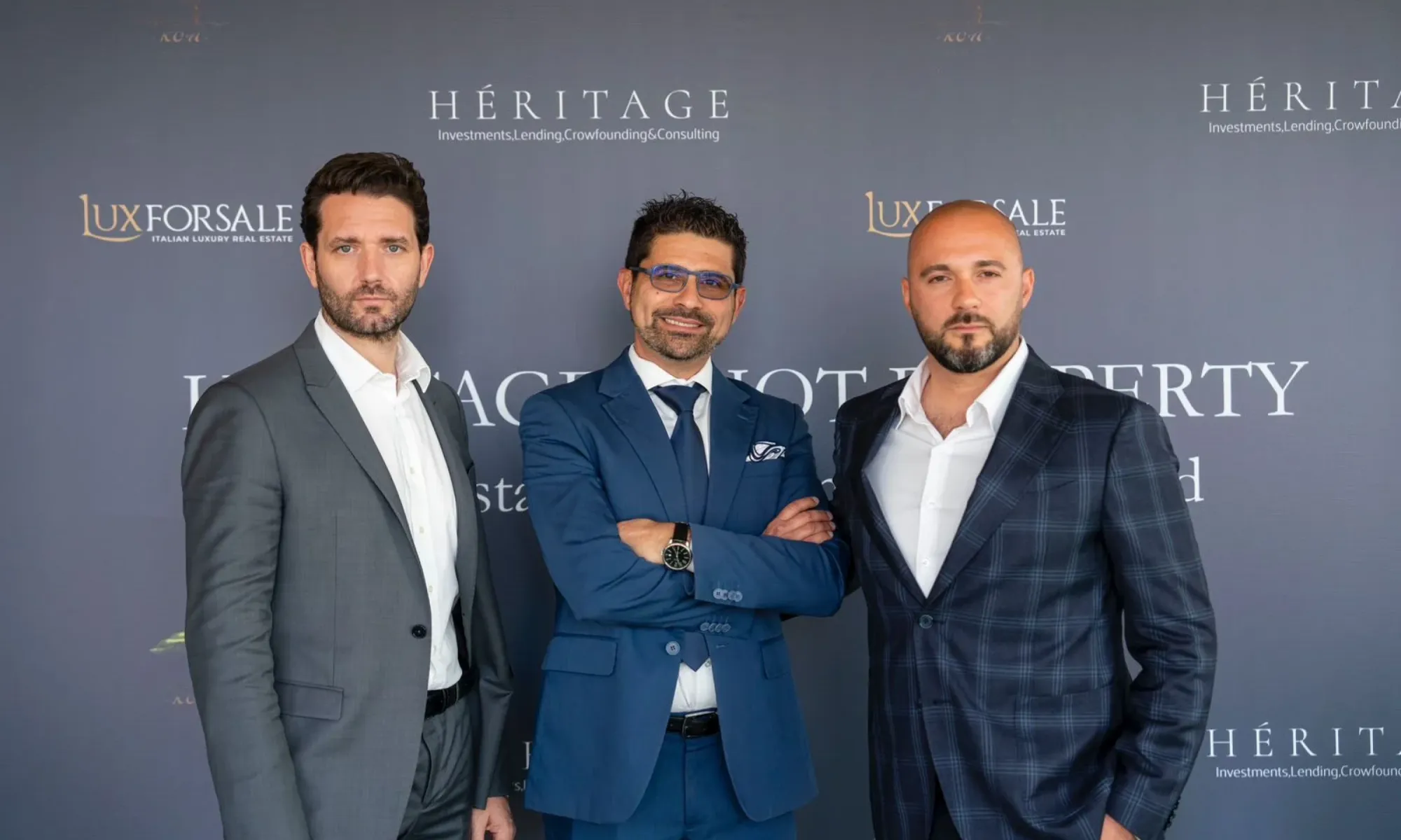 Three men in business suits pose for a photo at a Heritage Investments and Luxforsale real estate event backdrop.