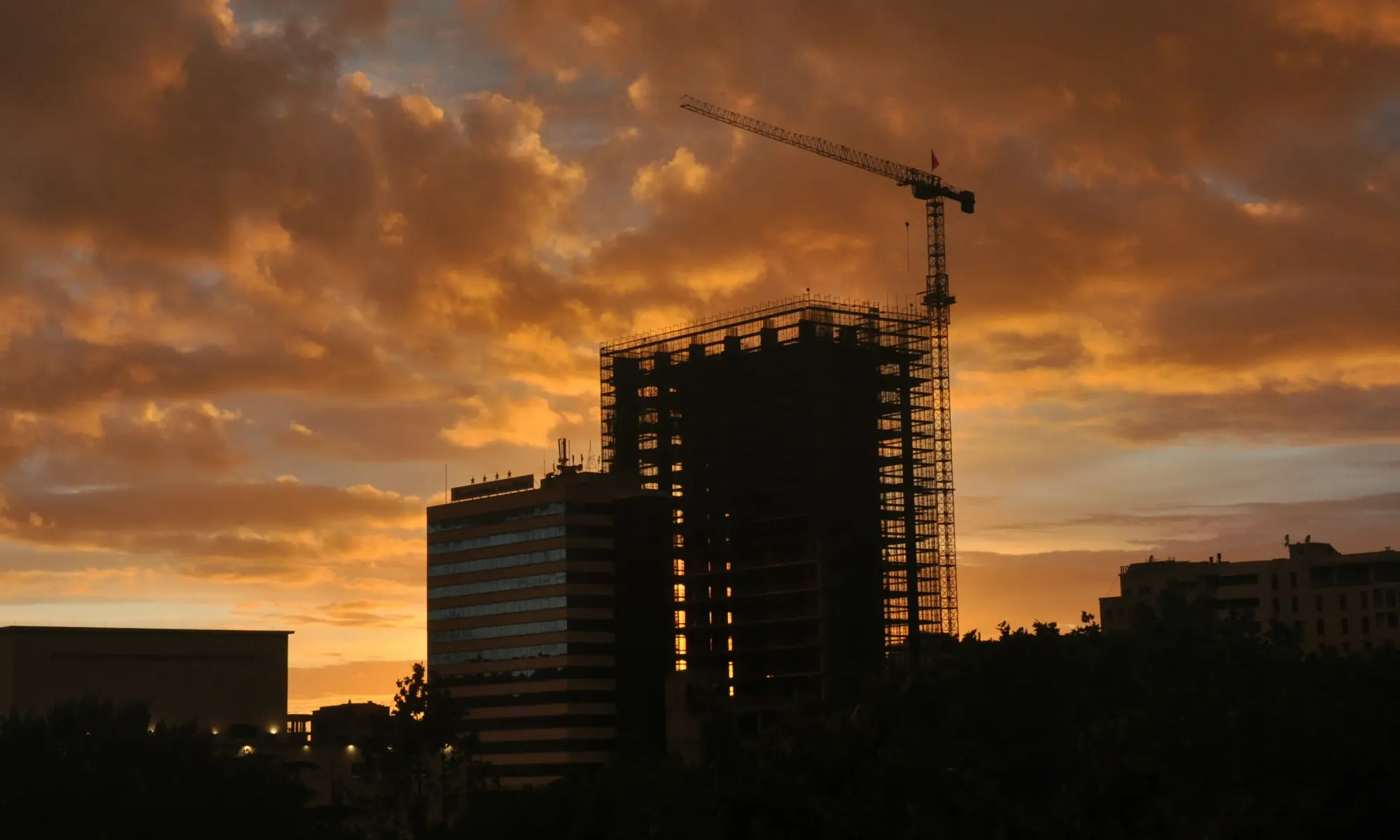 A high-rise building under construction with a crane silhouetted against a dramatic sunset sky.