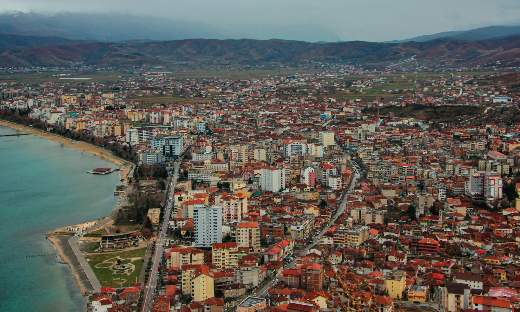 Aerial view of Pogradec, Albania, showing dense buildings with red rooftops along the lakeshore.