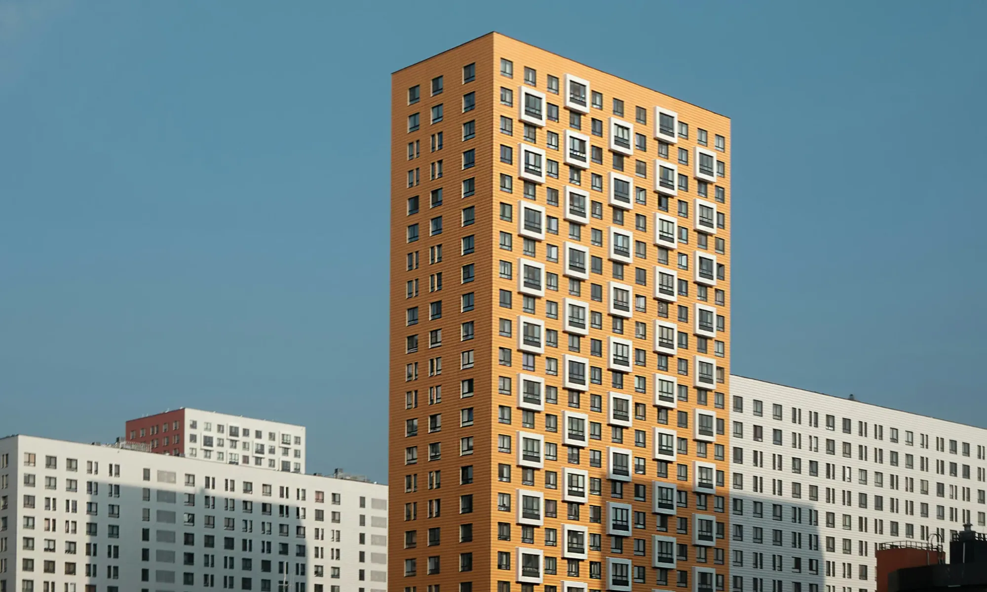 Tall orange residential high-rise with square windows, surrounded by white apartment blocks against a blue sky.