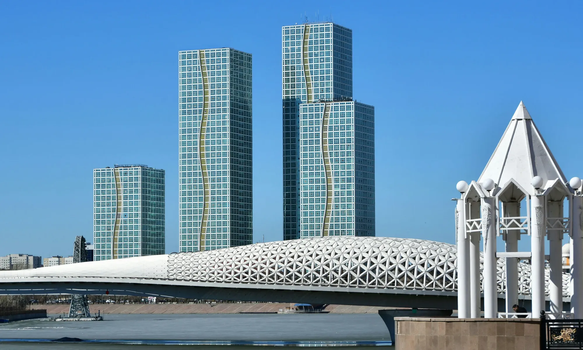 Modern high-rise towers with wavy gold accents behind a futuristic white bridge over a river in Astana under a clear blue sky.