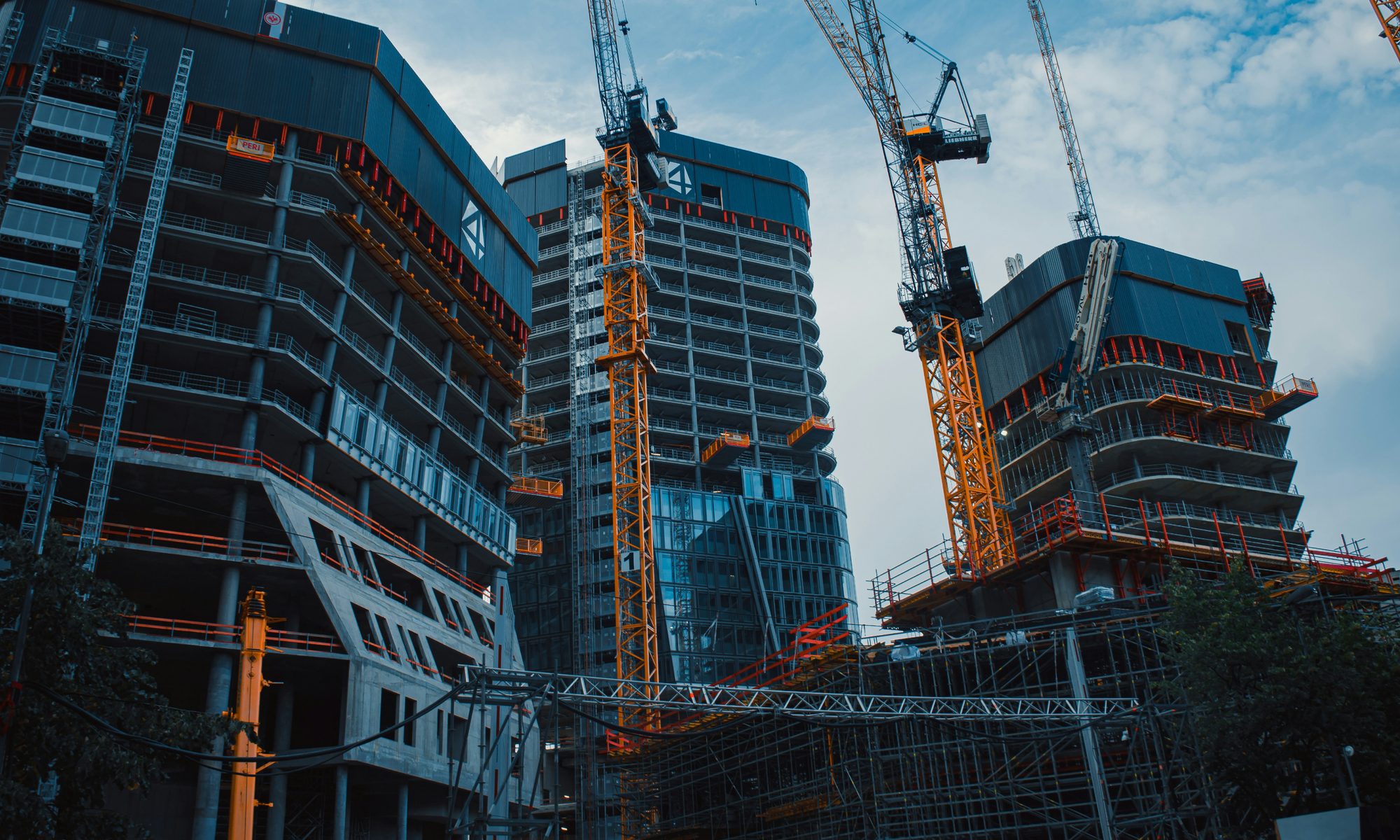 High-rise buildings under construction with multiple yellow cranes and scaffolding against a cloudy sky.