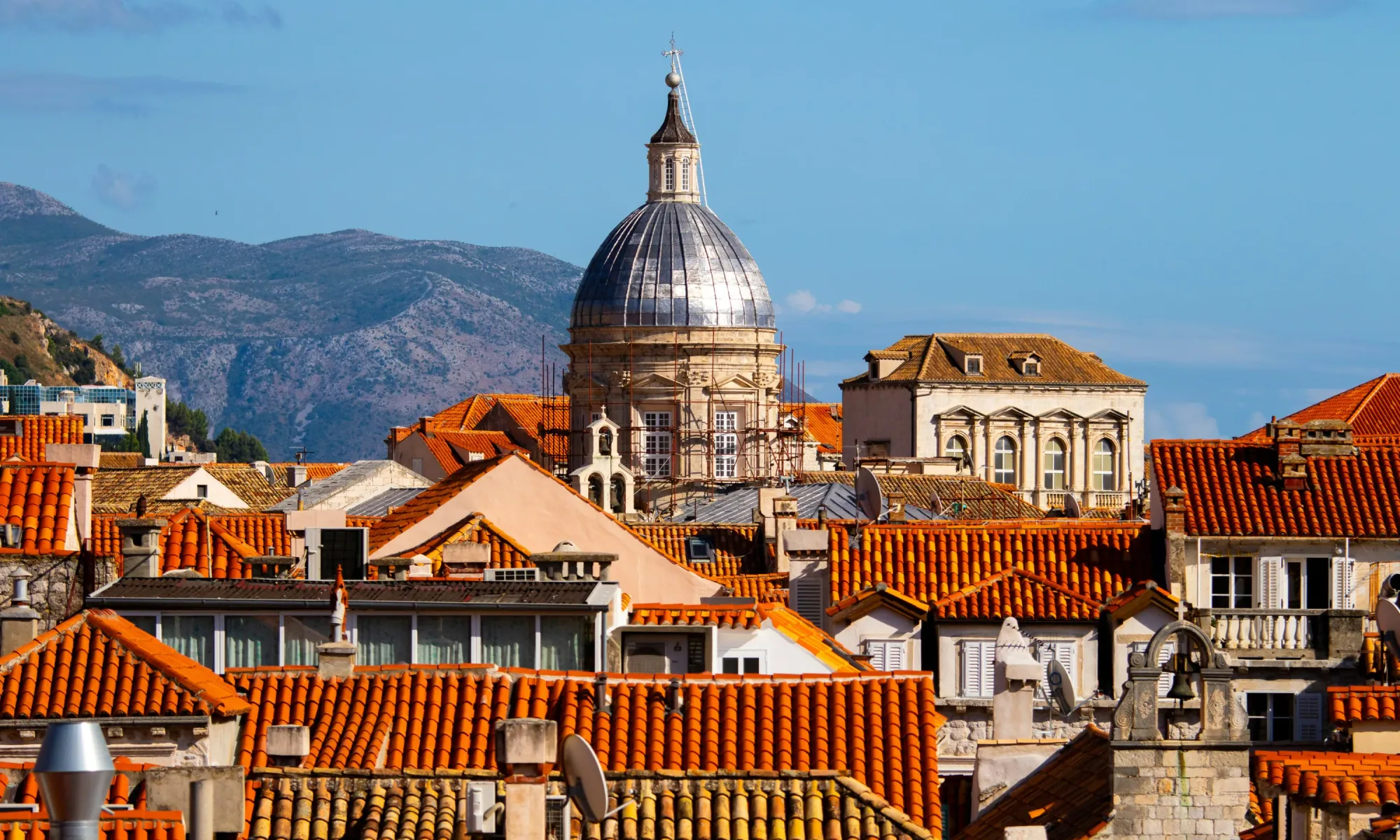 Close-up view of Dubrovnik’s cathedral dome rising above red-tiled rooftops with mountains in the background.