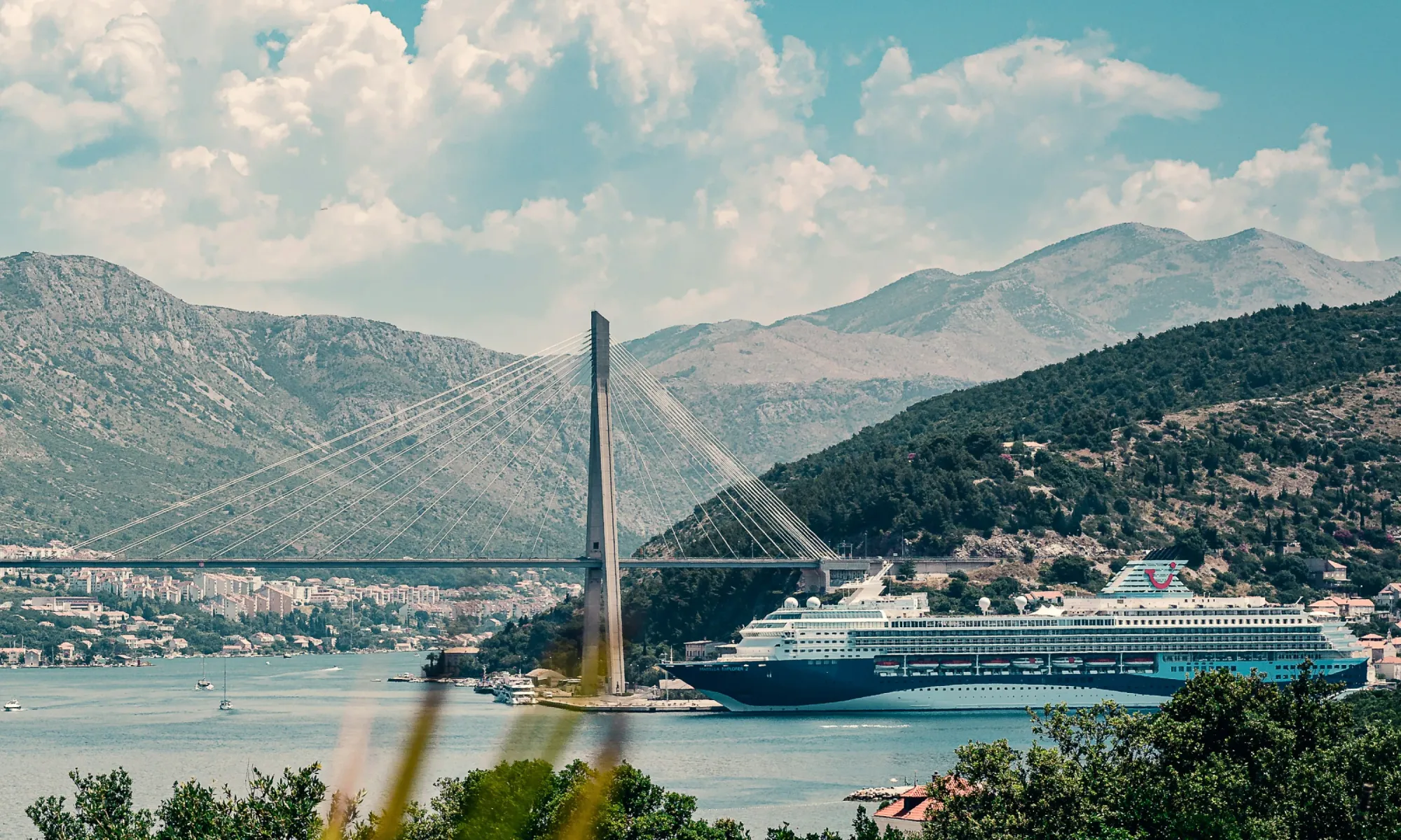Cable-stayed Franjo Tuđman Bridge in Dubrovnik with a large cruise ship docked below, mountains in the background.