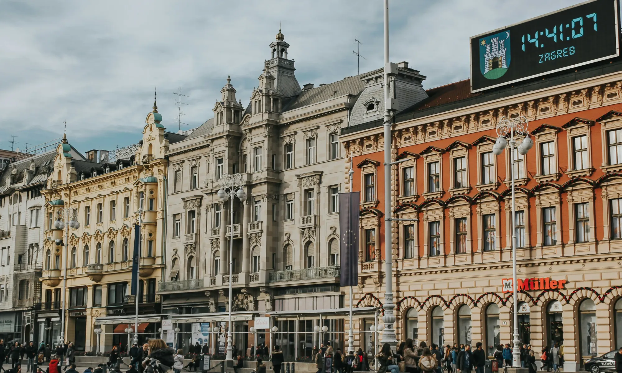 Historic buildings in Zagreb’s main square with ornate facades, a digital clock showing the city emblem, and people walking below.