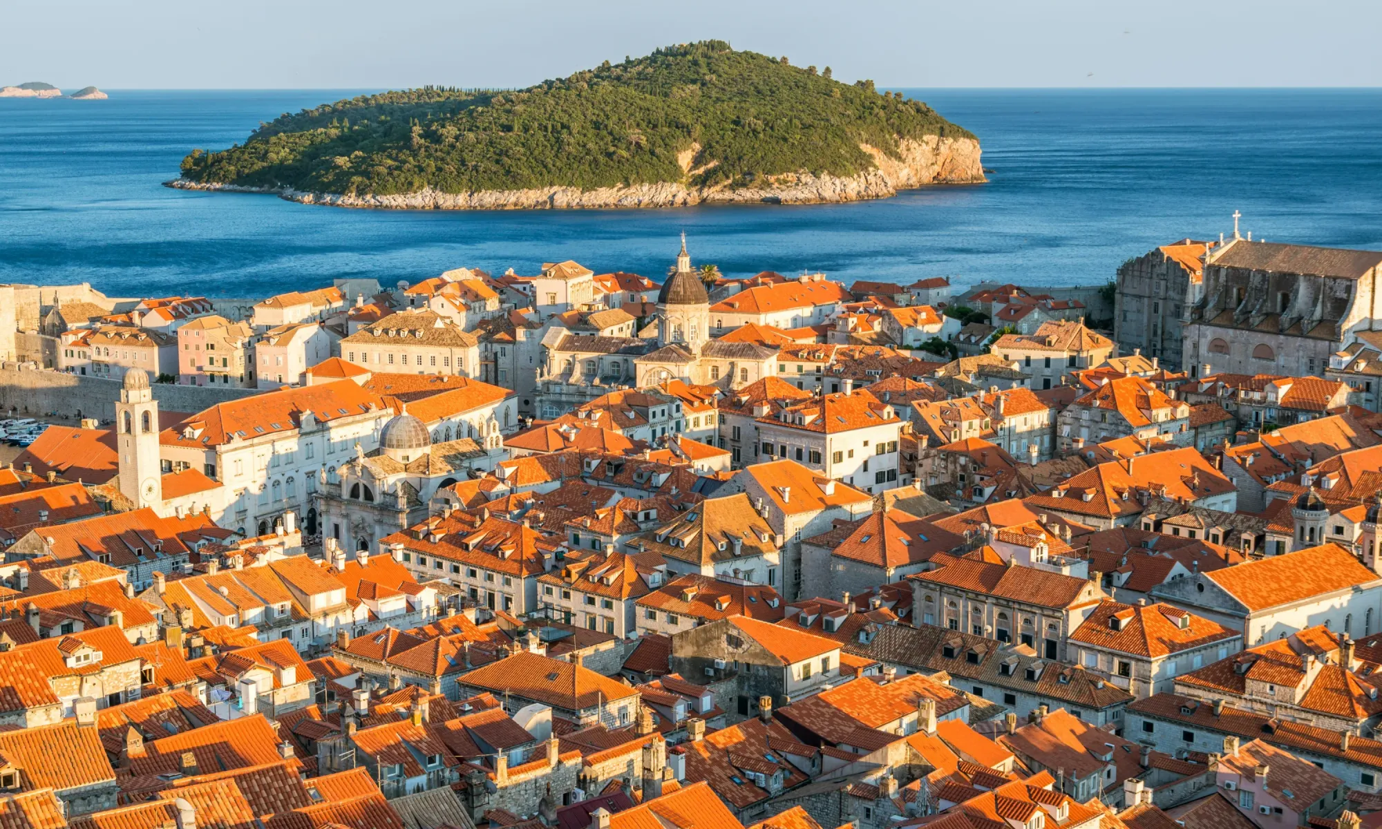 Panoramic view of Dubrovnik’s terracotta rooftops with a domed church and the Adriatic Sea, featuring Lokrum Island in the background.