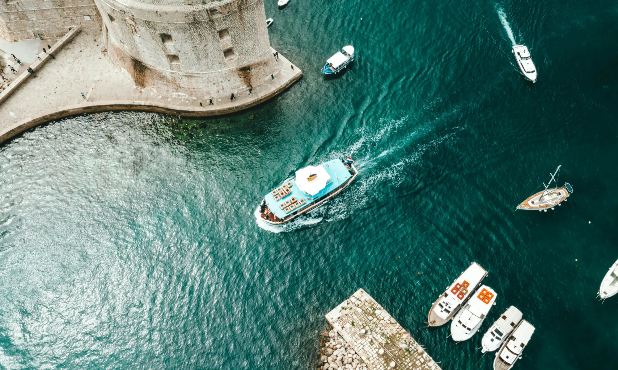 A tourist boat sails past the stone walls of Dubrovnik’s old fortress, surrounded by turquoise Adriatic waters and moored yachts.