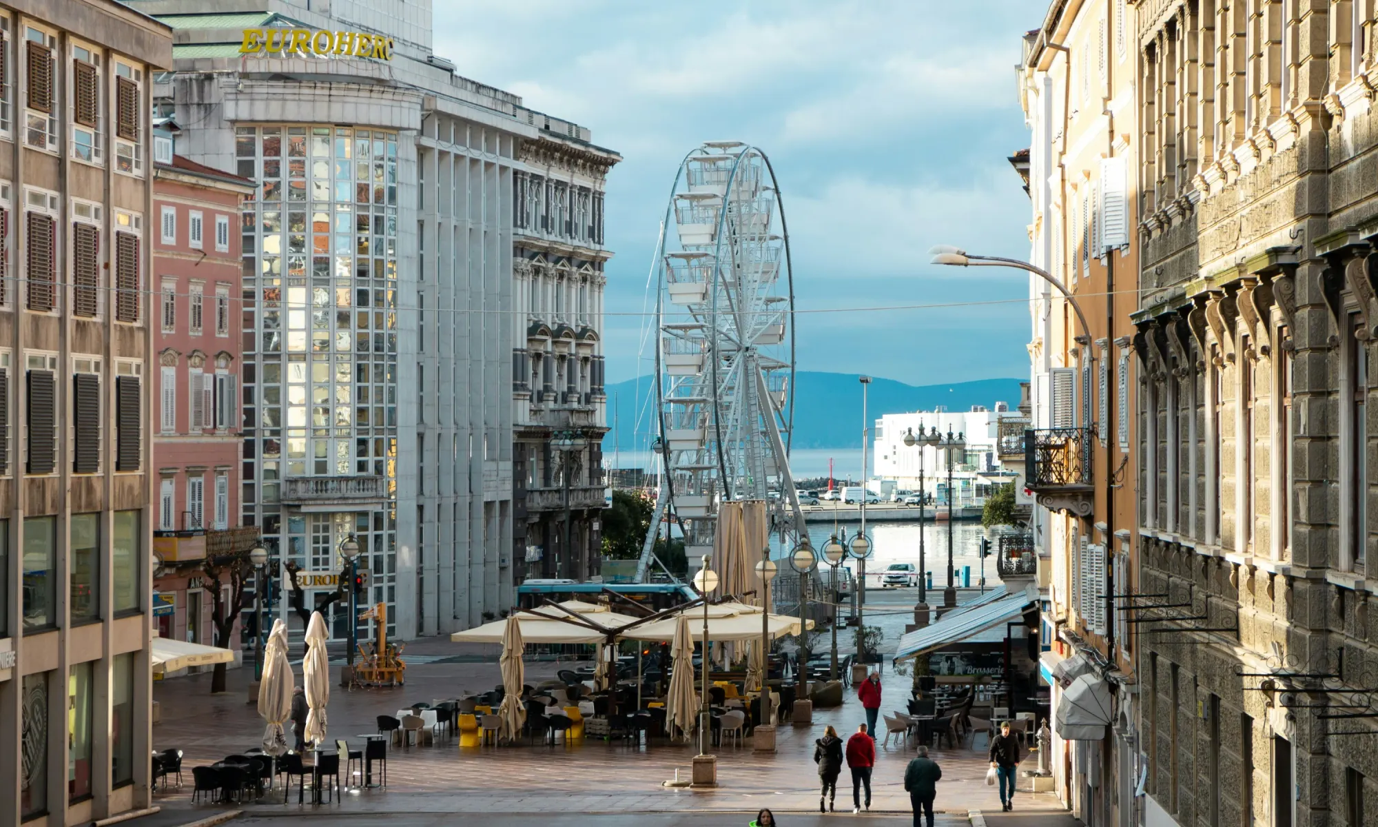 Urban street in Rijeka, Croatia, with outdoor cafes, historic buildings, and a large ferris wheel near the waterfront.
